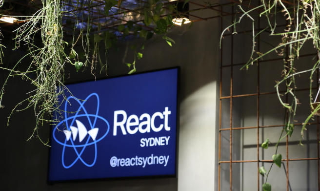 Photo of a wall mounted screen at a React Sydney event - displaying the React Sydney logo.